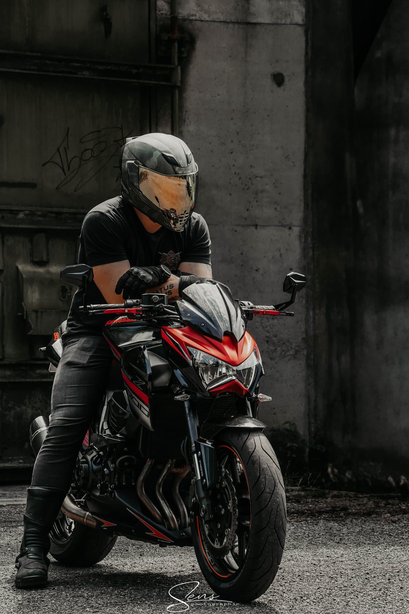 Motard casqué assis sur une moto rouge et noire devant un mur de béton