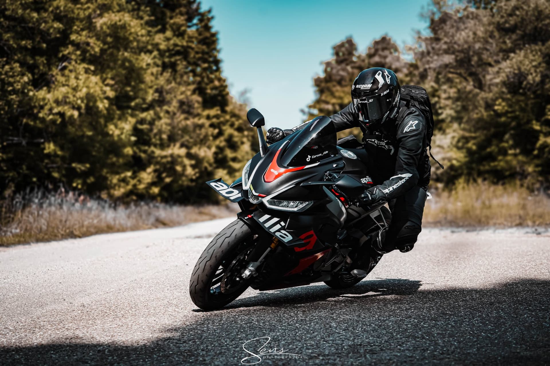 Motard penché dans un virage sur une route ensoleillée bordée d'arbres