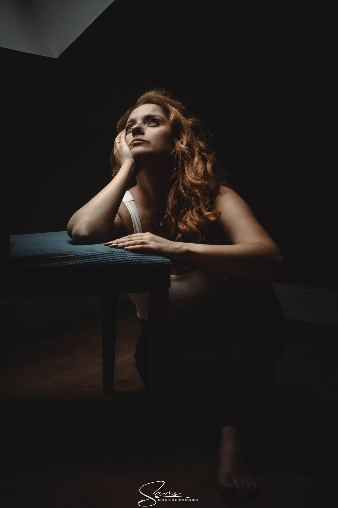 Femme aux cheveux roux accoudée à un tabouret, le regard levé vers une lucarne, dans une pièce sombre