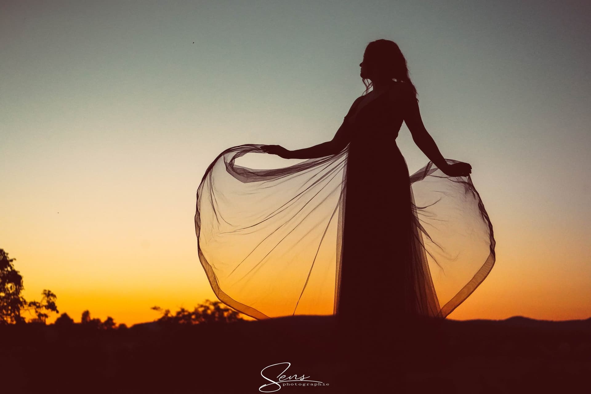 Silhouette d'une femme déployant une robe de tulle sur un ciel orangé de coucher de soleil