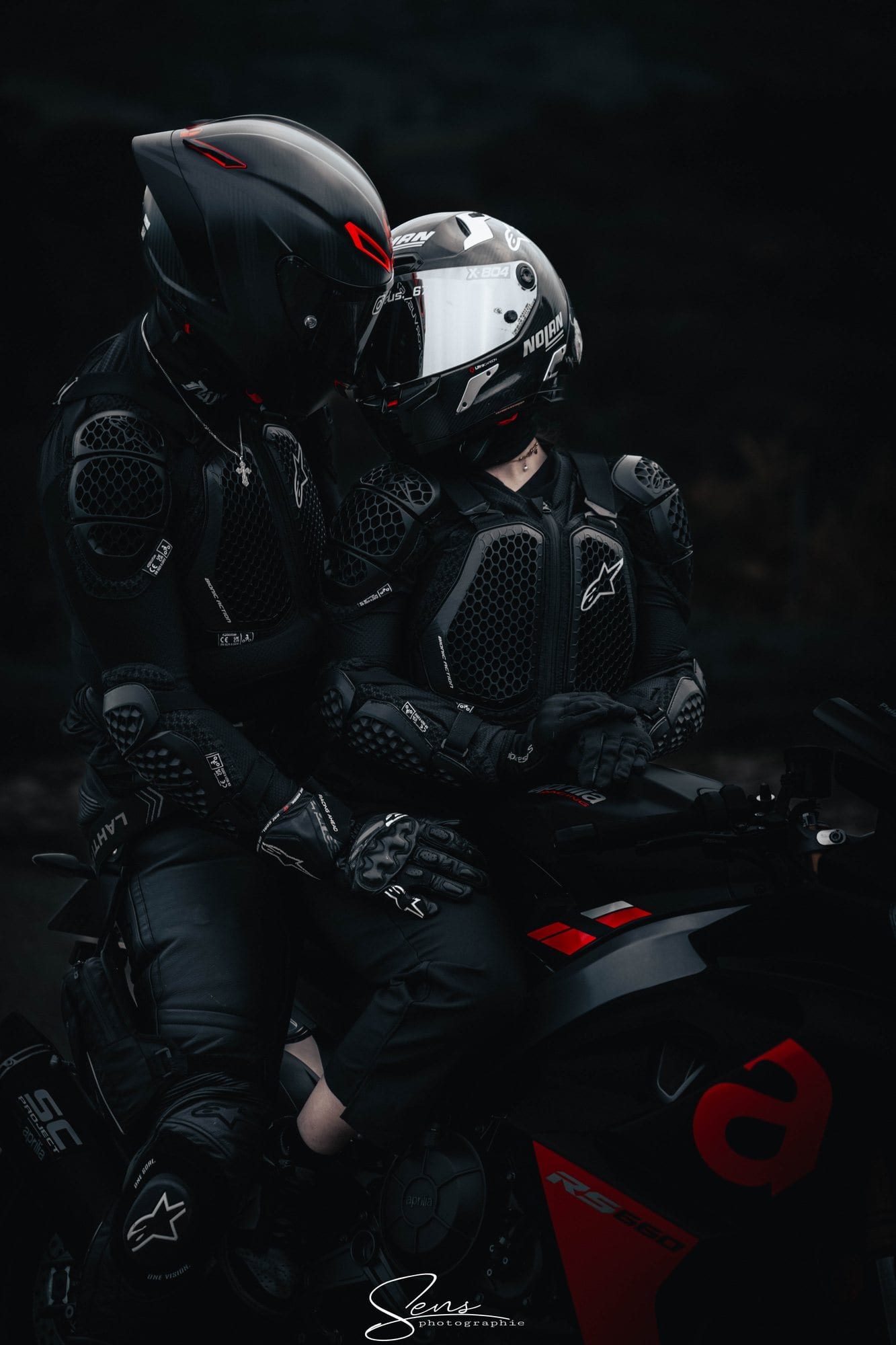 Deux motards casqués et en combinaison, front contre front sur une moto, dans la pénombre